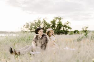 Couple sitting together in a sunlit meadow filled with tall grass and orange wildflowers, wearing hats and smiling. after